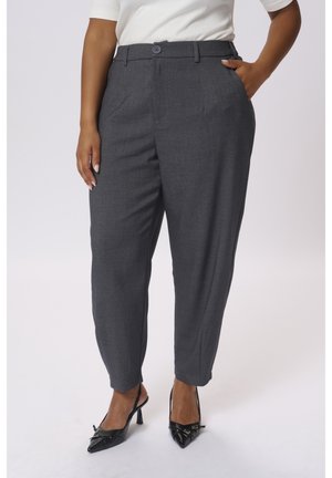 Persona con pantalones grises oscuros hechos a medida con botón y cremallera, combinados con tacones altos puntiagudos negros y una blusa blanca.