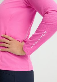 Långärmad skjorta i strukturerad rosa tyg med en figurnära design; har vit branding på ärmen. Modellen har en hand på höften.