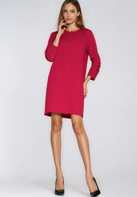 Kobieta stoi, ubrana w czerwoną sukienkę do kolan z długimi rękawami oraz czarne szpilki na jasnym szarym tle.