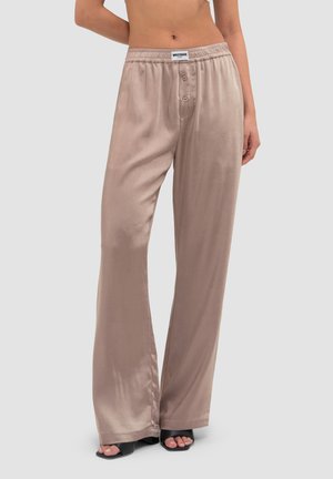 Frau trägt lockere, hoch taillierte beige Satinhose mit Knopfleiste und schwarze offene Sandalen mit Absatz auf grauem Hintergrund.