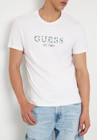 T-shirt blanc à manches courtes avec un logo "GUESS" à motifs verts et le texte "EST. 1981". Tissu en coton doux avec une coupe classique.