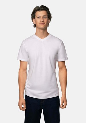 Camiseta blanca de cuello en V y mangas cortas, hecha de una tela suave. Llevada con jeans azul oscuro, el modelo se encuentra de pie contra un fondo neutro.