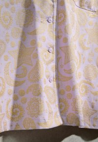 Tissu violet clair avec motifs célestes jaunes et motifs cachemire, comprenant des boutons et une petite poche sur le côté droit.