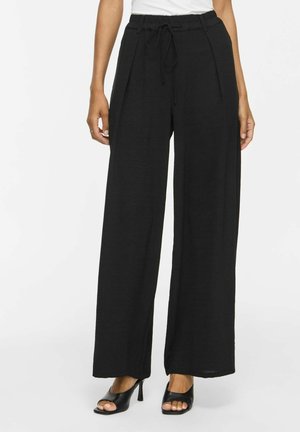 Femme portant un pantalon large noir ample avec un cordon à l'avant, associé à des chaussures noires à talons hauts ouverts sur fond blanc.