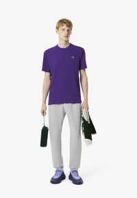 Camiseta de algodón morada con un logotipo verde, pantalones de chándal grises y zapatillas moradas. Los accesorios incluyen una bolsa verde y una toalla con estampado.