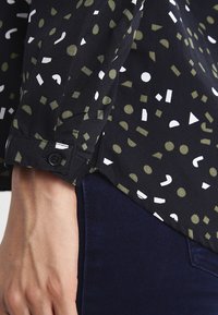 Camicia nera a maniche lunghe con varie forme geometriche in bianco e verde oliva. Dettaglio dei polsini abbottonati, tessuto leggero, motivo moderno.