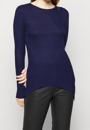 Frau trägt einen dunkelblauen, gerippten Pullover mit langen Ärmeln und asymmetrischem Saum sowie schwarze Hosen vor neutralem Hintergrund.