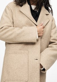 Abrigo de mezcla de lana beige con cuello de pico, cierre de un botón y bolsillo de parche delantero, que presenta una textura suave y un corte relajado.