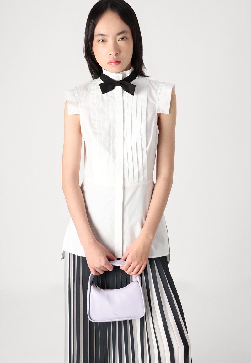 Blusa blanca con plisados y mangas cortas, acompañada de una corbata negra, combinada con una falda a rayas, sosteniendo un pequeño bolso de lavanda.