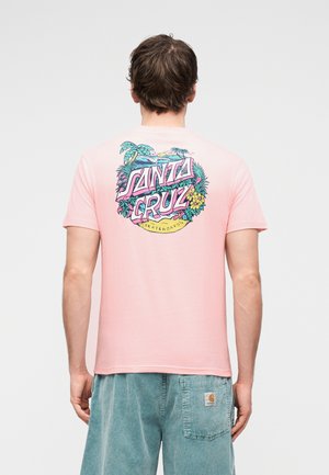 Férfi világos rózsaszín Santa Cruz pólóban, trópusi virágmintával és felirattal, valamint kopott kék rövidnadrágban, háttal áll egyszínű háttér előtt.