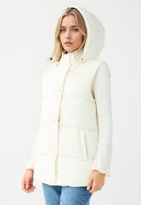 Weißer, plüschiger Weste mit Kapuze, vorderen Knöpfen, zwei vorderen Taschen und strukturiertem Stoff, getragen über einem hellen, langärmeligen Oberteil.