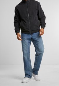 Man med svart zip-up jacka, blå jeans och vita sneakers som står mot en vanlig ljus bakgrund.