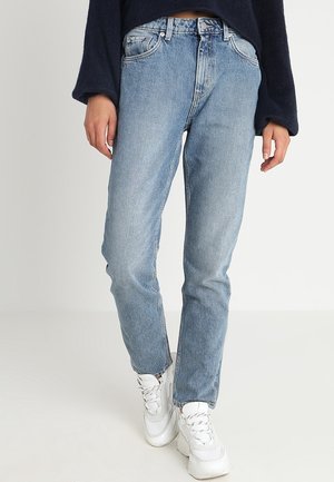 Person trägt hellblaue Jeans mit geradem Bein, ein dunkles Langarmshirt und weiße klobige Sneaker und steht auf einem einfarbigen Hintergrund.