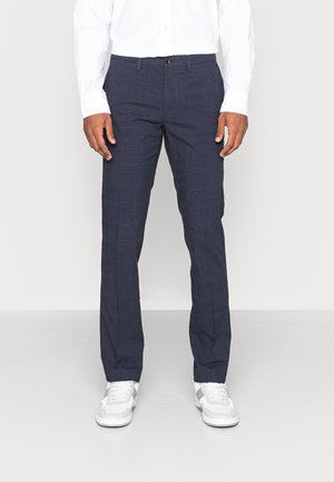 Uomo con pantaloni a quadri blu navy, camicia bianca a maniche lunghe e sneakers bianche, in piedi davanti a uno sfondo semplice.