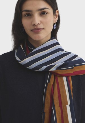 Femme aux cheveux foncés portant des boucles d'oreilles violettes en grappes et une écharpe rayée bleue, marron, rouge et beige sur un haut sombre.