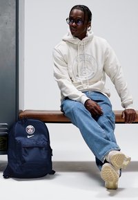 Mochila azul-marinho com logótipo de Paris e marca Nike, colocada num banco de madeira, ao lado de uma pessoa vestindo um hoodie creme e jeans azuis.