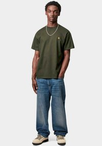 T-shirt verde oliva con colletto rotondo, abbinata a jeans blu larghi. Il modello è in una luce neutra, con una collana d'argento come accentuazione.