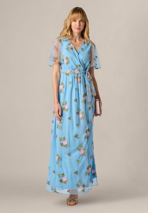 Kobieta w długiej, sięgającej podłogi, jasnoniebieskiej sukni z różowym kwiatowym haftem, przezroczystymi krótkimi rękawami, trzymająca beżową kopertówkę.