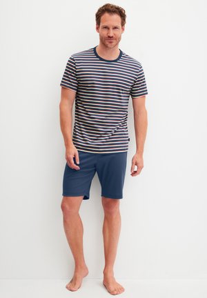 Mann steht barfuß vor weißem Hintergrund, trägt ein T-Shirt mit marineblauen, weißen und roten Streifen sowie einfarbige marineblaue Knie kurze Shorts.