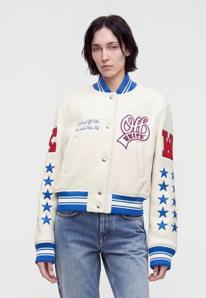 Person med kort mørkt hår iført hvid varsityjakke med blå stjerner og røde bogstaver, kombineret med blå jeans, stående mod hvid baggrund.