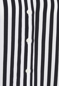 Camicia a righe verticali bianca e nera con una texture liscia, caratterizzata da una fila di quattro bottoni bianchi rotondi sul davanti.