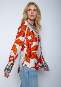 Jedwabna bluzka z dużymi pomarańczowymi wzorami kwiatowymi na jasnym tle. Posiada długie rękawy z czarnymi wzorzystymi mankietami oraz zakrzywiony dół.