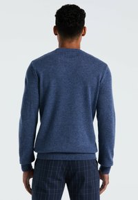 Man met kort krullend haar, gekleed in een blauwe gebreide trui en donkere blauwe geblokte broek, met zijn rug naar de camera gericht tegen een effen achtergrond.