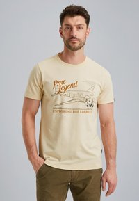 Beżowa koszulka bawełniana z krótkim rękawem, z grafiką samolotu i napisem "Pme Legend odkrywając żywioły" w brązowym druku.
