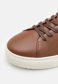 Sneaker in pelle marrone con finitura testurizzata, punta rotonda e suola in gomma bianca. Presenta lacci marroni e dettagli cuciti.