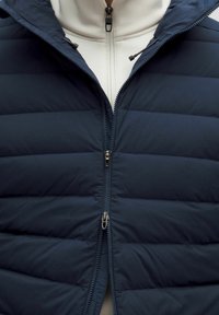 Tengerészeti pufi mellény vízszintes steppelt mintával, dupla cipzárral és rejtett zsebekkel, világos színű, cipzáros dzseki fölé rétegezve.