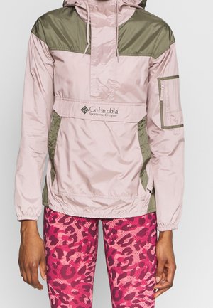 Veste coupe-vent Columbia Sportswear rose clair et olive avec poche kangourou à l'avant, demi-fermeture éclair et poche zippée en mesh sur la manche gauche, portée avec un legging camouflage rose.