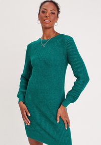 Vestido suéter verde de punto con un patrón texturizado, escote en V, mangas largas y puños acanalados, que ofrece una silueta ajustada.