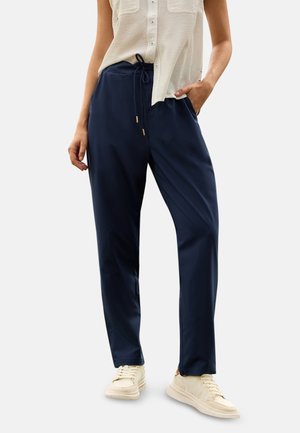 Persona vistiendo pantalones azul marino con cordón, top sin mangas color crema con botones y zapatillas crema, de pie con una mano en el bolsillo.