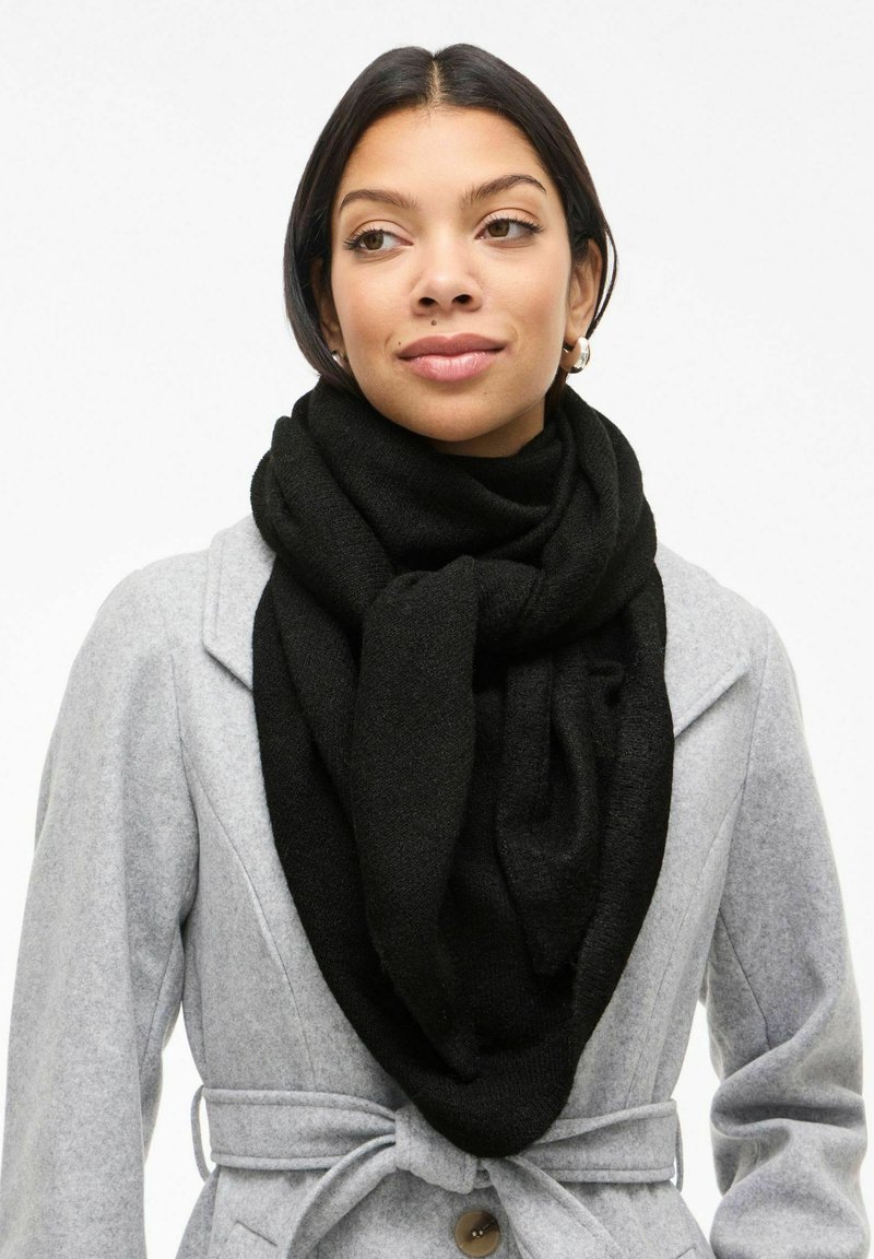 Bufanda de punto negra con una textura suave, colocada de manera suelta alrededor del cuello. Lleva puesta sobre un abrigo gris claro con cintura atada.