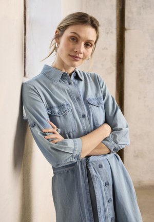 Kobieta w jasnoniebieskiej sukience dżinsowej z podwiniętymi rękawami, z założonymi rękami, opierająca się o fakturowaną, kremową ścianę wewnątrz pomieszczenia.