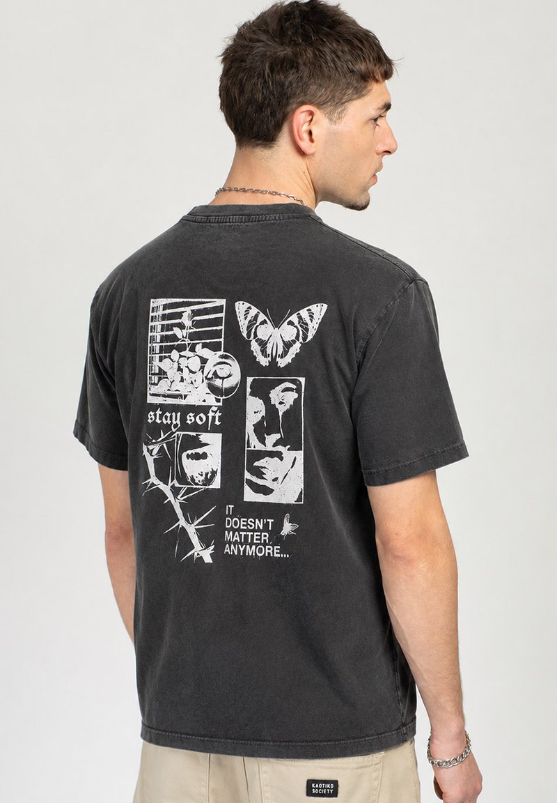 Άντρας που κοιτάει πλαγίως φορώντας μαύρο T-shirt με λευκή γραφιστική τέχνη πεταλούδας, προσώπων, αγκαθιών και κείμενο που γράφει "stay soft" και "it doesn't matter anymore."