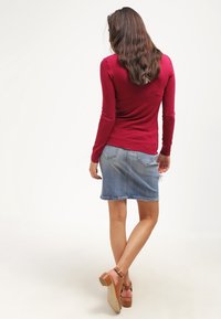 Top rosso a maniche lunghe abbinato a una gonna di jeans blu, con dettagli alle tasche posteriori e tacchi spessi marroni con cinturini alla caviglia.