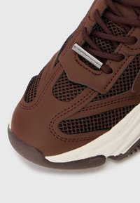 Baskets brunes avec des panneaux en maille, dotées d'un bout en caoutchouc lisse, d'une tige en synthétique et d'une étiquette logo argentée sur la zone de laçage.