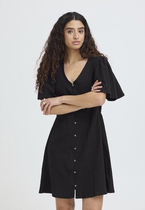 Mujer con cabello largo y rizado, vestida con un vestido negro abotonado con mangas abullonadas, de pie con los brazos cruzados frente a un fondo liso.