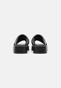 Sandales mules en cuir noir avec une semelle épaisse. Dotées de larges brides croisées et d'une texture lisse, conçues pour un style minimaliste.