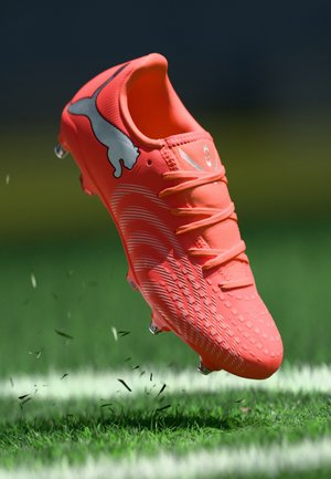 Crampon de football orange vif avec surface texturée, accents blancs et design à lacets, flottant au-dessus d'une herbe verte et des marques blanches du terrain.