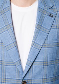 Niebieska marynarka w kratę z żółtymi i białymi akcentami, z klapami w kształcie notcha, zapięciem na jeden guzik i czarną przypinką na klapie. Noszona na białym t-shircie.