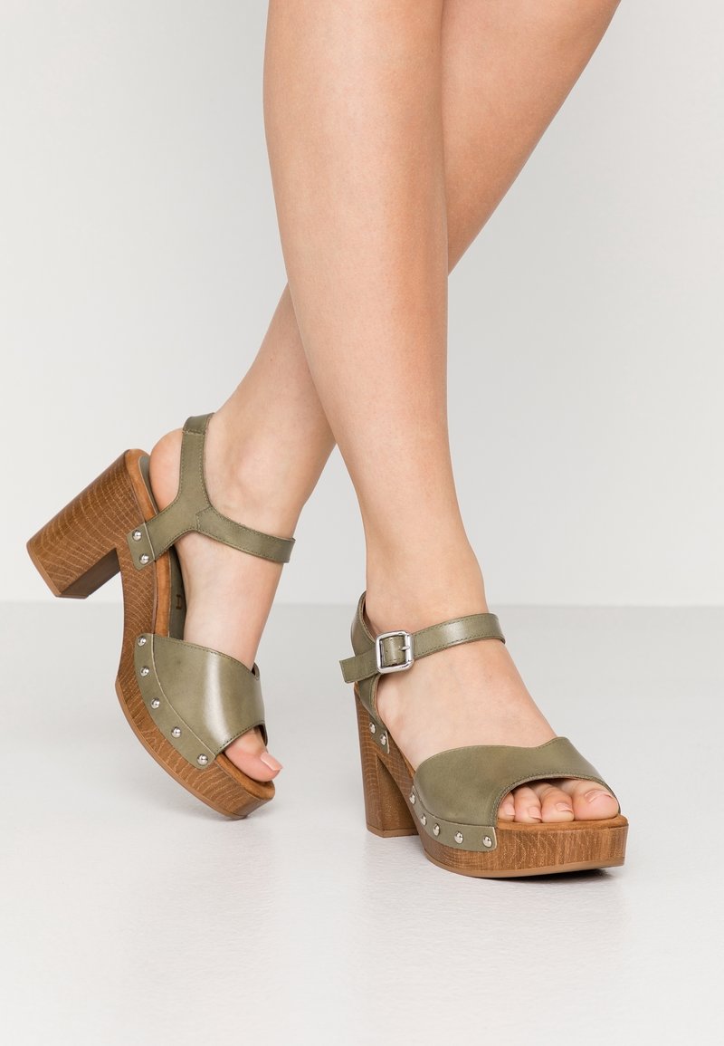 Jambes féminines portant des sandales à plateforme vert olive ouvertes à l'avant, avec des talons épais en bois et des brides à la cheville ornées de clous métalliques.