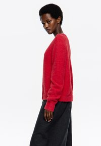 Suéter rojo, esponjoso, con mangas largas, escote redondo y un corte relajado. Combinado con pantalones de pierna ancha en gris oscuro.