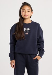 Niña joven con cabello largo y oscuro que lleva una sudadera azul marino con "1989 Estado de Nueva York" y una bandera, pantalones azules marinos a juego, de pie con una mano en el bolsillo.