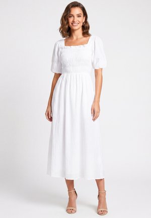 Donna sorridente che indossa un vestito midi bianco con maniche corte a sbuffo e scollo quadrato, abbinato a tacchi aperti beige, in piedi su sfondo bianco.