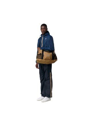 Uomo con tuta blu navy con strisce gialle e scarpe da ginnastica bianche che tiene una borsa da viaggio beige e nera con dettaglio del logo.