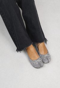 Pantofole ballet in suede grigio con punta rotonda, dettaglio decorativo a fiocco e leggere variazioni di colore, abbinate a jeans neri di denim sfilacciati.