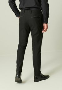 Pantalones negros a medida con corte ajustado, con elegantes bolsillos traseros con detalles en negro, y combinados con zapatos de vestir negros brillantes.