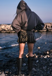 Un sweat à capuche gris surdimensionné, une jupe évasée tie-dye verte et des bottes en caoutchouc noires jusqu'aux genoux. La scène présente un rivage rocheux avec de l'eau en arrière-plan.